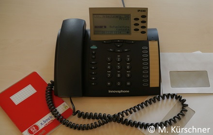 Ein schwarzes Tastentelefon mit Display. Links daneben ein rotes Schreibheft, rechts danaben zwei weisse Briefumschläge.