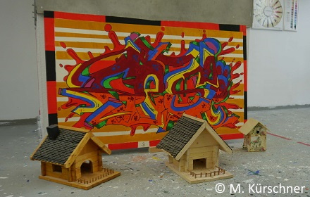 Im Hintergrund ein buntes Bild. Im Vordergrund der unterschiedlich große Vogelhäuschen aus Holz.
