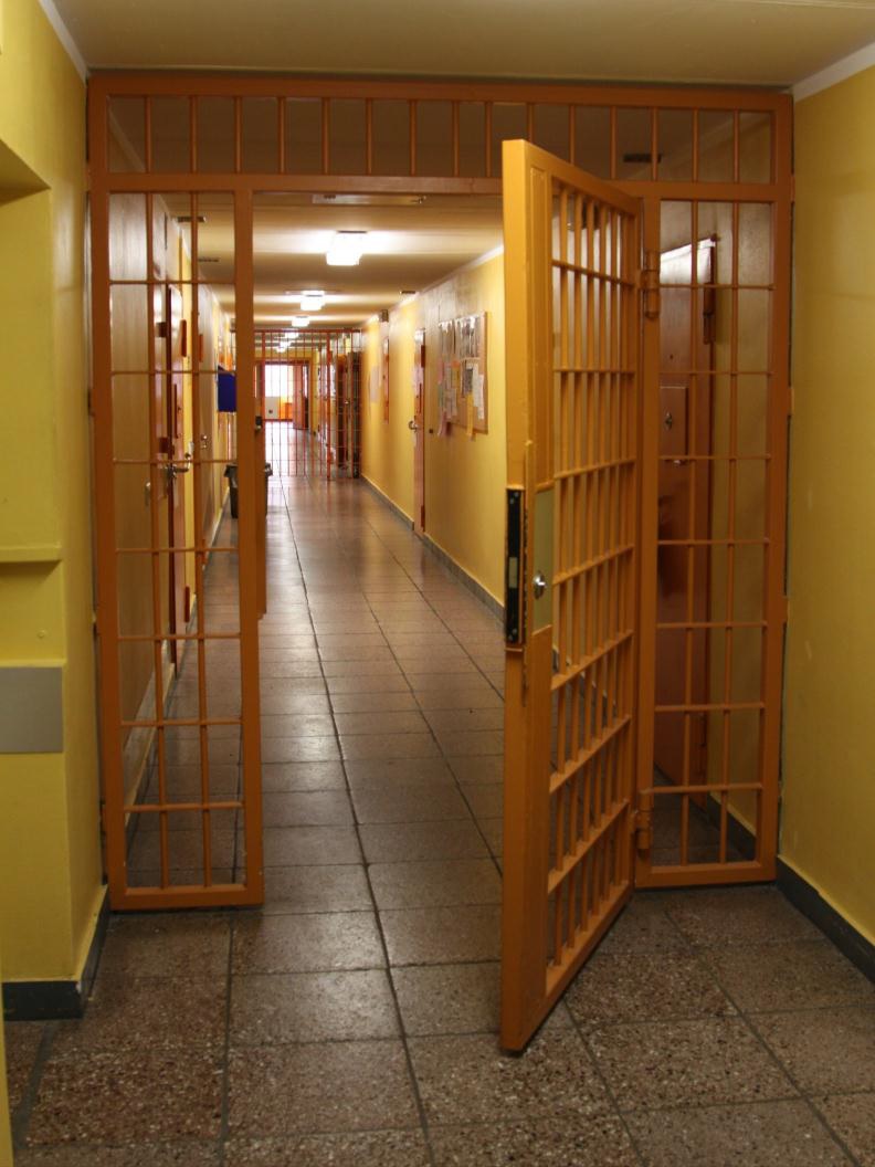 Hafthaus Ein langer Flur mit gelber Wandfarbe, dazwischen mehrere Ebenen, die durch Gittertüren, die gelb lackiert sind, getrennt werden. Eine der Gittertüren ist geöffnet, die im hinteren Bereich ist geschlossen.