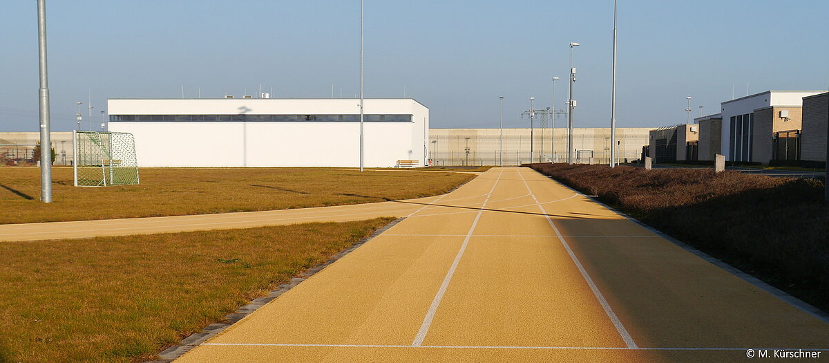 Blick auf die Sporthalle und den Sportplatz einem Fußballtor und der 100 Meter Strecke. Rechts die Ausbildungsbetriebe. Im Hintergrund die Anstaltsmauer.