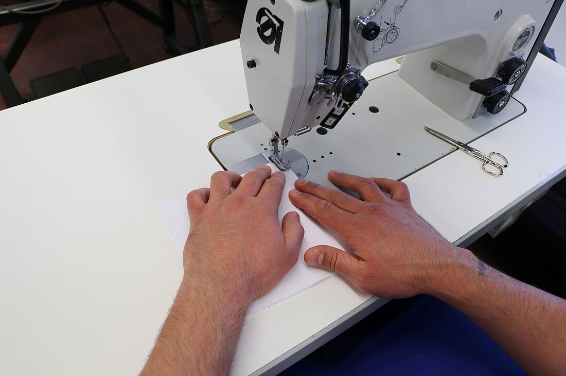 ein Mann sitzt an einer Nähmaschine und bereitet diese für das Nähen vor. Neben seiner rechten Hand liegt eine kleine silberne Schere. Die Nähmaschine ist grau und ist in einen weißen Tisch eingelassen.