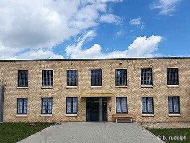 Das Foto zeigt die Arrestanstalt in der Frontalansicht. Man blickt genau auf die Eingangstür und die sich links und rechts anschließenden vergitterten Fenster. Vor der Tür ist noch ein Stück des gepflasterten Zugangsweges zu sehen. Über dem Gebäude sieht man einen blauen Himmel mit vielen kleinen weißen Wolken.