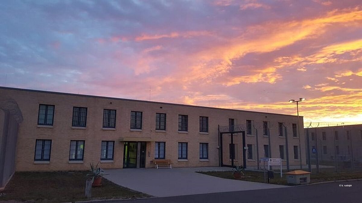 Das Foto zeigt das Gebäude des Jugendarrest in der Frontaufnahme. Zu sehen ist mittig die Eingangstür und links und rechts daneben sowie oberhalb der Tür sind die Fenster mit der typischen Vollzugsvergitterung zu sehen. Am rechten Bildrand sieht man ebenfalls ein Tor aus groben Stahlmaschen auf dessen oberem Rand Stacheldraht angebracht ist. Zusätzlich ist einkleines Stück des Zufahrtsweges hinter dem Tor zu sehen. Vor der Tür, links und rechts der Zuwegung stehen zwei Blumenkübel mit Agarven. Ebenfalls sieht man einen Behälter mit Streugut, einen Abfalleimer und ein Schild mit der Aufschrift Thüringer Jugendarrestanstalt. Im Hintergrund ist ein, durch den Sonnenaufgang rosa, gelb, blau gefärbter Himmel zu sehen.