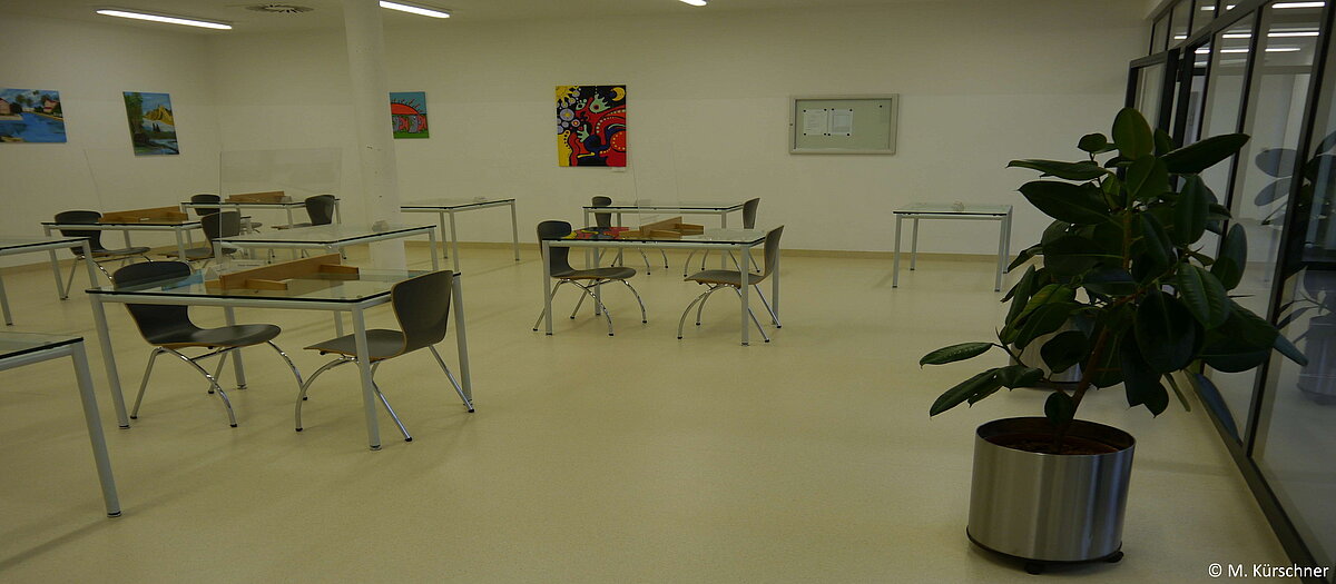 Besuchsbereich der Jugendstrafanstalt mit Glastischen, Stühlen, Bildern an der Wand und einer großen Pflanze.