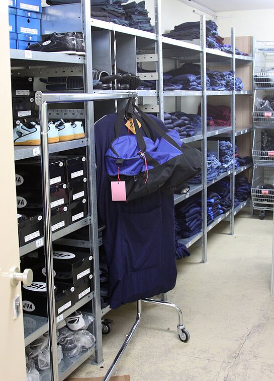 Kammer Die blaue Anstaltskleidung der Gefangenen lagert in der Kleiderkammer auf großen Regalen, die wiederum in mehrere Etagen aufgeteilt sind. Sodass ein Bereich für Lagerung von weißen Turnschuhen, mit gelben Sohlen vorgesehen ist. Daneben befindet sich ein rollbarer Kleiderständer, der eine blaue Sporttasche trägt. Dahinter werden zusammengelegte Hosen und Oberteile in jeweiligen Regalebenen gelagert.