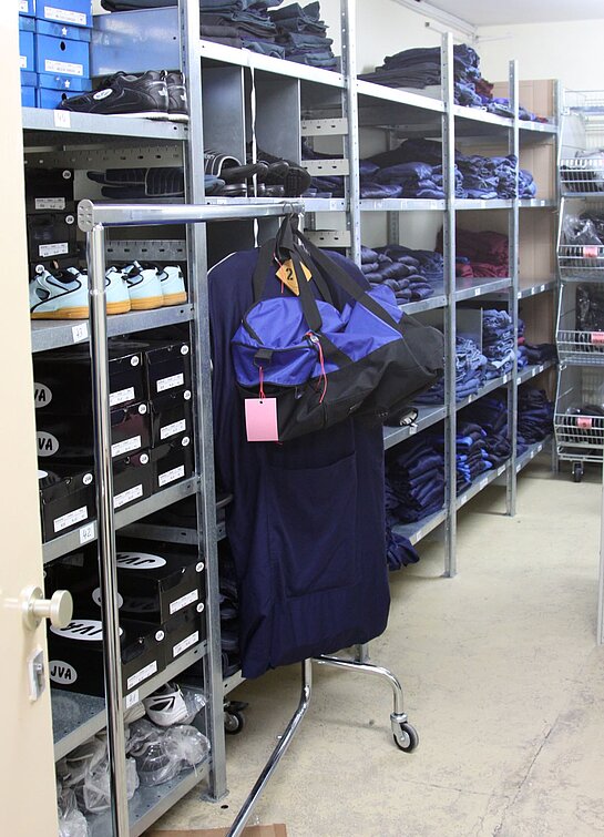 Die blaue Anstaltskleidung der Gefangenen lagert in der Kleiderkammer auf großen Regalen, die wiederum in mehrere Etagen aufgeteilt sind. Sodass ein Bereich für Lagerung von weißen Turnschuhen, mit gelben Sohlen vorgesehen ist. Daneben befindet sich ein rollbarer Kleiderständer, der eine blaue Sporttasche trägt. Dahinter werden zusammengelegte Hosen und Oberteile in jeweiligen Regalebenen gelagert.