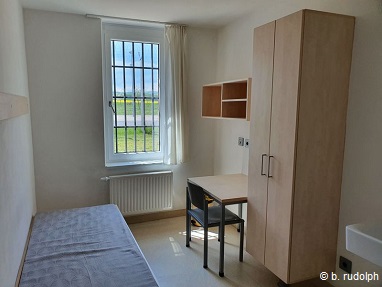 Das Foto zeigt einen Arrestraum der von der Tür aus fotografiert wurde. Auf der rechten Seite ist das Bett zu sehen, welches aus hellem Holz mit einer einfachen Matratze besteht. An der gegenüberliegenden Wand von der Tür aus gesehen befindet sich ein großes vergittertes Fenster durch das man einen schönen Blick auf die Rapsfelder und die Natur hat. In der rechten hinteren Ecke des Raumes steht ein heller Holztisch und ein Stuhl. Daran schließt sich ein zweitüriger Kleiderschrank an. Ganz links im Bild ist noch das Waschbecken zu sehen. Neben dem Waschbecken befindet sich die Tür hinter der die Toilette versteckt ist. Der Raum ist ca. 12 m² groß und hell und freundlich gehalten.