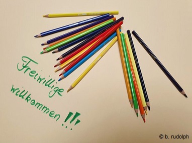 Das Bild zeigt auf einem cremefarbenen Untergrund eine grüne Schreibschrift mit den Worten Freiwillige willkommen. Über dem Schriftzug liegen wie zufällig verteilt achtzehn Buntstifte in verschiedenen Farben.