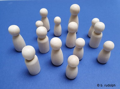 Das Foto zeigt vierzehn unterschiedlich große Holzfiguren. Diese Figuren sind ohne Arme und Beine und ohne Gesichter. Sie sind sehr einfach und schlicht gehalten und symbolisieren in ihrer unterschiedlichen Größe die unterschiedlichen Menschen die hier arbeiten. Der Hintergrund ist in einem meerblau gestaltet.