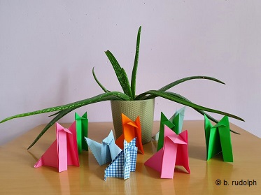 Auf dem Foto steht zentral ein heller Blumentopf mit einer ausladenden Agarve. Um die Pflanze herum stehen acht aus Papier gefaltete Origami Füchse in verschiedenen Farben. Drei davon sind in einem satten grün, zwei in knalligem Pink, einer in orange, zwei in hellblau und einer in einem weiß blau kariertem Muster.